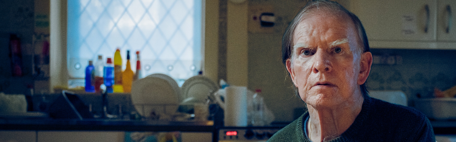 An older man sits in his kitchen, looking into the camera sadly