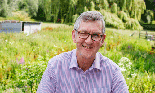 Keith Oliver, author of Dear Dementia, smiles at the camera with a green garden and wild flowers behind him