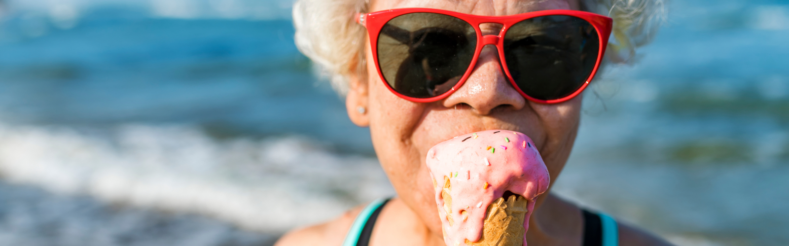 An older lady with sunglasses eats an ice cream by the sea
