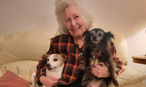 An older lady sits smiling at the camera with a dog under each arm