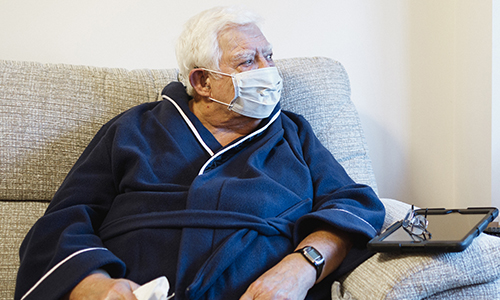 An older man, sat on a sofa and wearing a protective face mask