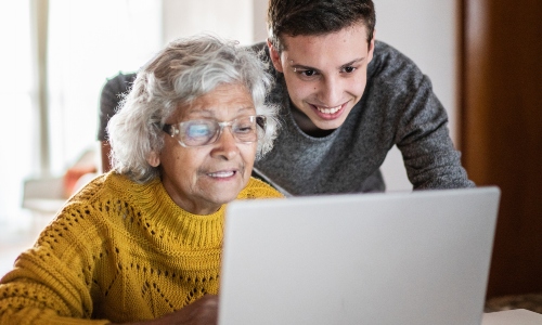 An older woman with glasses uses her laptop, while a younger man helps her