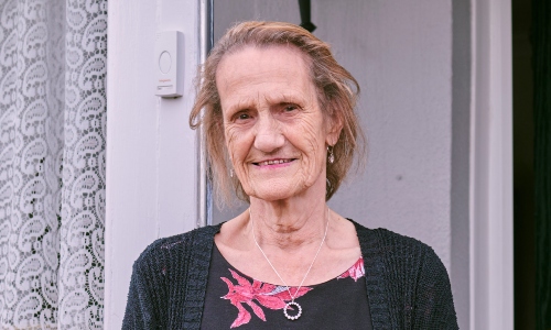 An older lady with a dark blonde bob smiles at the camera outside the front door of her house