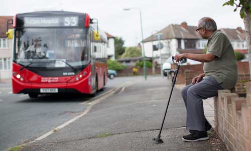 Older man sitting on a wall, holding his walking stick waiting for the bus to arrive